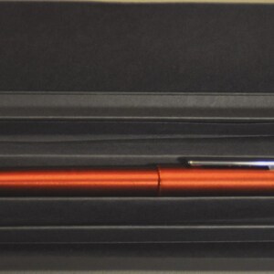 May include: A metallic orange fountain pen with a silver-colored clip rests horizontally inside a rectangular black presentation box. The pen features a smooth, cylindrical body and a matching cap, securely nestled within the dark, structured interior.