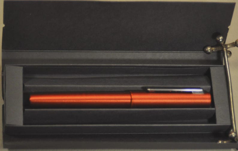 May include: A metallic orange fountain pen with a silver-colored clip rests horizontally inside a rectangular black presentation box. The pen features a smooth, cylindrical body and a matching cap, securely nestled within the dark, structured interior.