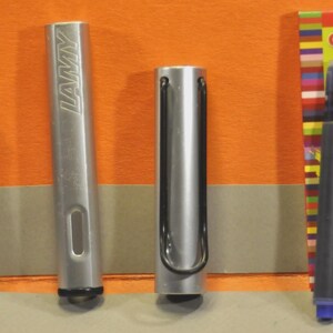 K&ouml;nnte beinhalten: Ein silberner Lamy-F&uuml;llhalter, zerlegt in Federteil, Schaft und Kappe mit schwarzem Drahtclip, dazu eine blaue Tintenpatrone und eine rote Schachtel mit der Aufschrift Universal-Tintenpatronen, die f&uuml;r Lamy- und Standard-F&uuml;llhalter geeignet ist.