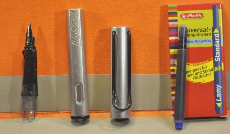 K&ouml;nnte beinhalten: Ein silberner Lamy-F&uuml;llhalter, zerlegt in Federteil, Schaft und Kappe mit schwarzem Drahtclip, dazu eine blaue Tintenpatrone und eine rote Schachtel mit der Aufschrift Universal-Tintenpatronen, die f&uuml;r Lamy- und Standard-F&uuml;llhalter geeignet ist.
