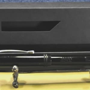 Puede incluir: Una pluma estilogr&aacute;fica negra brillante con un clip y un anillo de tapa en tono plateado, apoyada sobre un peque&ntilde;o soporte de metal frente a una caja de presentaci&oacute;n rectangular negra. La pluma presenta un grabado sutil en el cuerpo.