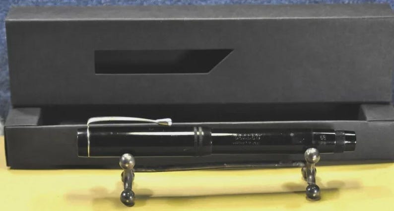 Puede incluir: Una pluma estilogr&aacute;fica negra brillante con un clip y un anillo de tapa en tono plateado, apoyada sobre un peque&ntilde;o soporte de metal frente a una caja de presentaci&oacute;n rectangular negra. La pluma presenta un grabado sutil en el cuerpo.
