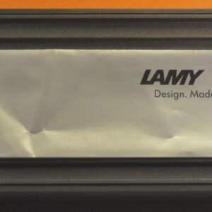 May include: A rectangular black box with a silver foil insert. The foil has the word "LAMY" printed in black, along with the text "Design. Made in Germany."