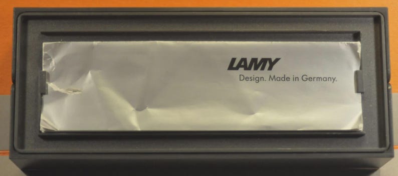 May include: A rectangular black box with a silver foil insert. The foil has the word "LAMY" printed in black, along with the text "Design. Made in Germany."