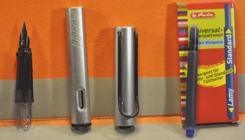 K&ouml;nnte beinhalten: Ein silberner Lamy-F&uuml;llhalter, zerlegt in Federteil, Schaft und Kappe, neben einer blauen Tintenpatrone und einer Schachtel Herlitz Universal-Patronen mit dem Text Standard, Lamy und K&ouml;nigsblau.
