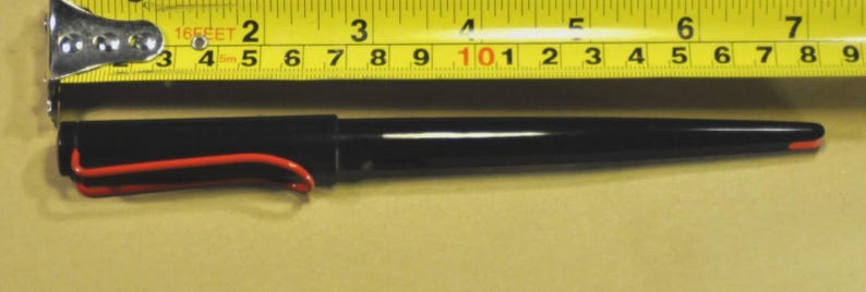 K&ouml;nnte beinhalten: Ein schwarzer Stift mit rotem Clip und Spitze, etwa 15 cm lang, liegt neben einem gelben Ma&szlig;band. Das Band zeigt Messungen in Fu&szlig; und Zoll an. Der Stift hat ein schlankes, modernes Design.