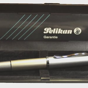 Puede incluir: Un bolígrafo plateado dentro de un estuche negro abierto. El bolígrafo tiene un clip metálico brillante y una punta redondeada. El interior del estuche es negro, con una tarjeta que muestra el logotipo de Pelikan y la palabra "Garantie".
