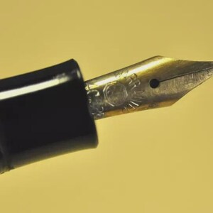 Puede incluir: Un plum&iacute;n de pluma estilogr&aacute;fica de metal dorado con un orificio de ventilaci&oacute;n central y texto grabado, unido a una secci&oacute;n de agarre de pl&aacute;stico negro brillante. El plum&iacute;n presenta una punta afilada y una hendidura central detallada.