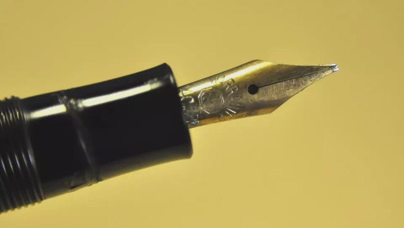 Puede incluir: Un plum&iacute;n de pluma estilogr&aacute;fica de metal dorado con un orificio de ventilaci&oacute;n central y texto grabado, unido a una secci&oacute;n de agarre de pl&aacute;stico negro brillante. El plum&iacute;n presenta una punta afilada y una hendidura central detallada.