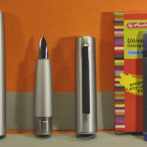 May include: A silver fountain pen with a black nib, disassembled, next to a box of blue ink cartridges. The box is red with colorful stripes and text that reads "Universal-tintenpatronen". A blue ink cartridge is also visible.