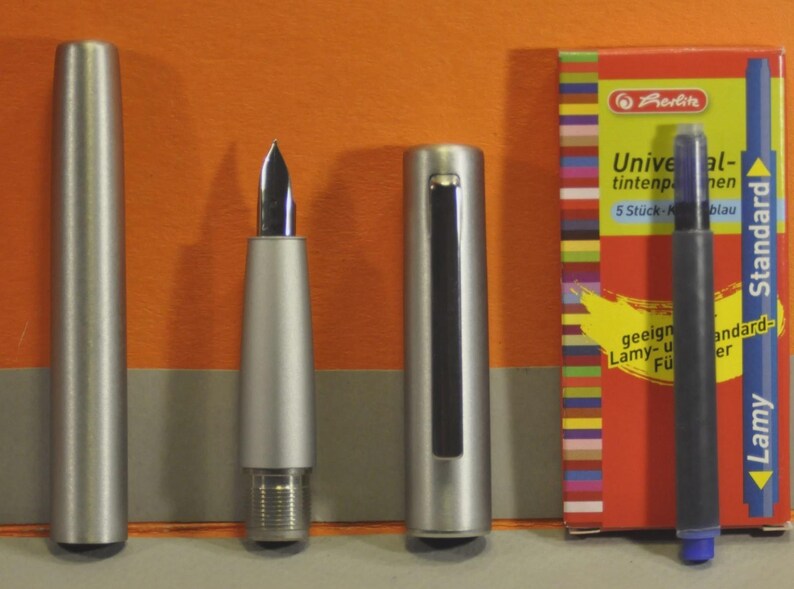 May include: A silver fountain pen with a black nib, disassembled, next to a box of blue ink cartridges. The box is red with colorful stripes and text that reads "Universal-tintenpatronen". A blue ink cartridge is also visible.
