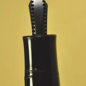 Puede incluir: Un plum&iacute;n de pluma estilogr&aacute;fica negro con su secci&oacute;n de agarre. El plum&iacute;n met&aacute;lico presenta una punta afilada y una ranura central, extendi&eacute;ndose desde una empu&ntilde;adura cil&iacute;ndrica de pl&aacute;stico negro. La secci&oacute;n incluye finas estr&iacute;as horizontales.