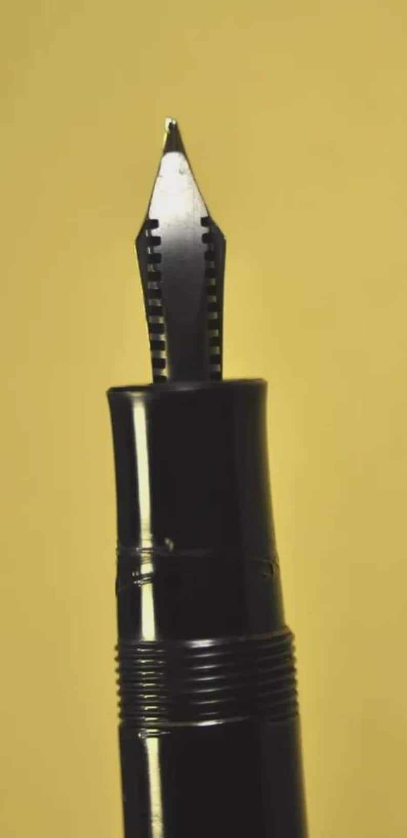 Puede incluir: Un plum&iacute;n de pluma estilogr&aacute;fica negro con su secci&oacute;n de agarre. El plum&iacute;n met&aacute;lico presenta una punta afilada y una ranura central, extendi&eacute;ndose desde una empu&ntilde;adura cil&iacute;ndrica de pl&aacute;stico negro. La secci&oacute;n incluye finas estr&iacute;as horizontales.