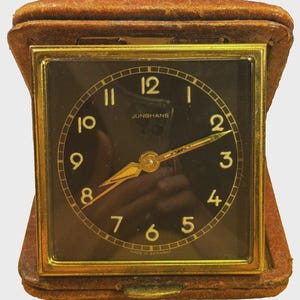 May include: A square Junghans travel alarm clock with a black face, gold-colored numerals, and gold-colored hands. The clock is housed in a brown leather folding case with a gold-colored metal frame surrounding the glass clock face.