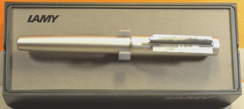 May include: A silver-colored Lamy pen rests on a gray box. The pen has a cylindrical body and a rectangular clip. The box has the Lamy logo in black on the top left corner. The pen is secured by two gray plastic holders.