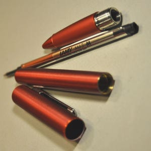 May include: A disassembled metallic orange pen consisting of a conical cap, a cylindrical barrel, and a separate ink refill cartridge. The refill is labeled LAMY M63 M. The pen components feature a smooth, matte finish with silver-toned metal accents.