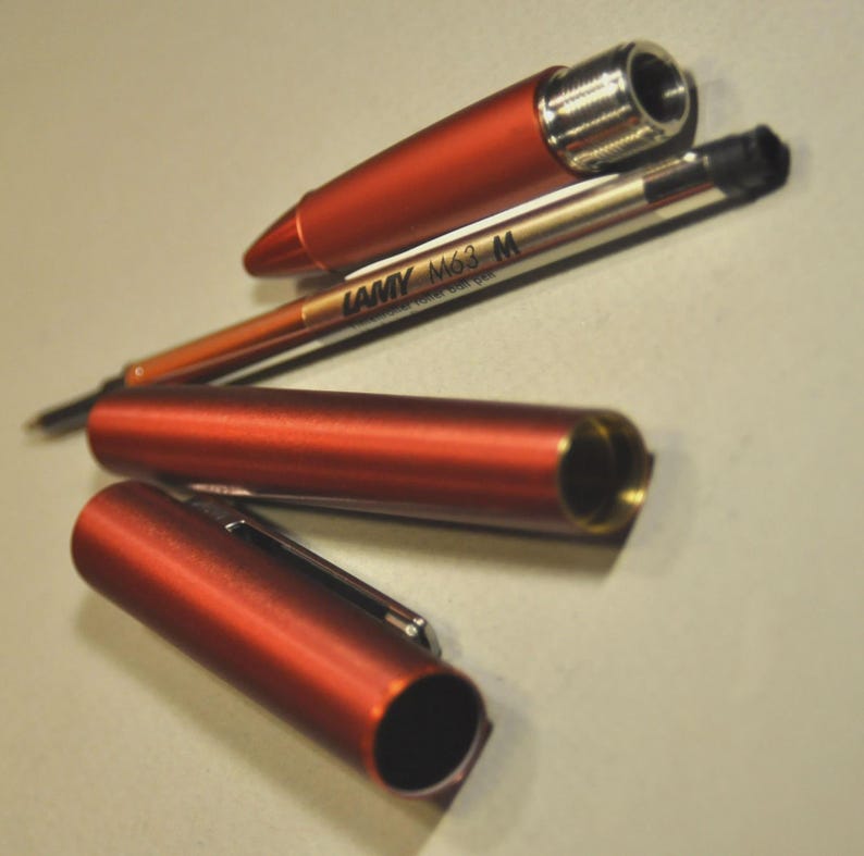 May include: A disassembled metallic orange pen consisting of a conical cap, a cylindrical barrel, and a separate ink refill cartridge. The refill is labeled LAMY M63 M. The pen components feature a smooth, matte finish with silver-toned metal accents.