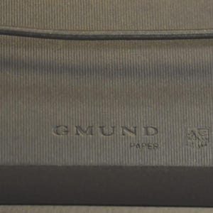 May include: A dark gray, textured paper surface featuring the embossed text GMUND PAPER and a small, square-shaped emblem. A thin, dark horizontal cord or string runs across the upper portion of the frame, resting against the ribbed material.
