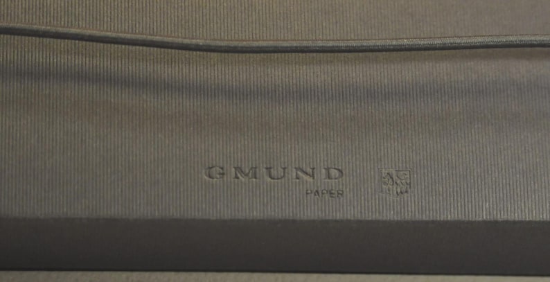 May include: A dark gray, textured paper surface featuring the embossed text GMUND PAPER and a small, square-shaped emblem. A thin, dark horizontal cord or string runs across the upper portion of the frame, resting against the ribbed material.