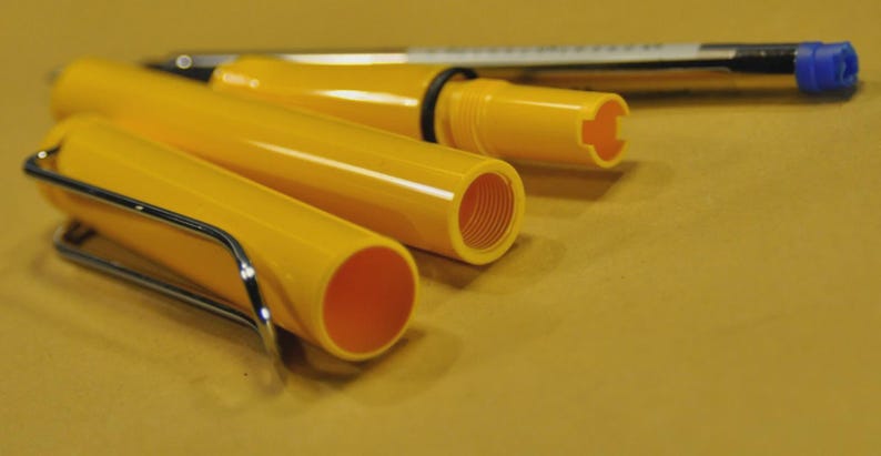 Puede incluir: Un bol&iacute;grafo amarillo desmontado con un clip plateado y un bol&iacute;grafo de tinta negra con una tapa azul. Las piezas del bol&iacute;grafo est&aacute;n dispuestas sobre una superficie amarilla, mostrando los componentes internos del instrumento de escritura. El dise&ntilde;o del bol&iacute;grafo es simple y funcional.