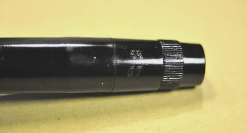 Puede incluir: Un bol&iacute;grafo o instrumento de escritura cil&iacute;ndrico de color negro brillante con una secci&oacute;n de agarre texturizada cerca del extremo. El cuerpo muestra los n&uacute;meros 55 y 07 grabados verticalmente en el lateral, con un acabado liso.