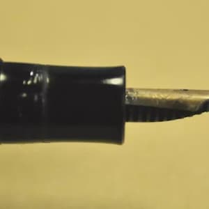 Puede incluir: Una pluma estilogr&aacute;fica negra con un plum&iacute;n met&aacute;lico de tono plateado. El cuerpo de la pluma tiene un acabado liso y brillante con una secci&oacute;n estriada cerca de la zona de agarre, que conduce al plum&iacute;n met&aacute;lico expuesto que se extiende horizontalmente.