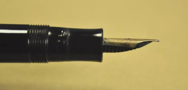 Puede incluir: Una pluma estilogr&aacute;fica negra con un plum&iacute;n met&aacute;lico de tono plateado. El cuerpo de la pluma tiene un acabado liso y brillante con una secci&oacute;n estriada cerca de la zona de agarre, que conduce al plum&iacute;n met&aacute;lico expuesto que se extiende horizontalmente.