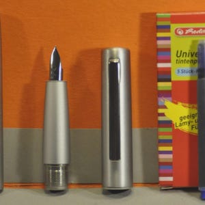May include: A disassembled silver fountain pen with a nib, cap, and barrel, alongside a box of blue ink cartridges. The box is red with colorful stripes and text that reads "Universal tintenpatronen". A blue ink cartridge is also visible.
