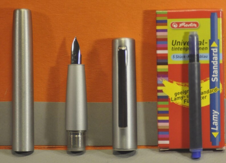 May include: A disassembled silver fountain pen with a nib, cap, and barrel, alongside a box of blue ink cartridges. The box is red with colorful stripes and text that reads "Universal tintenpatronen". A blue ink cartridge is also visible.