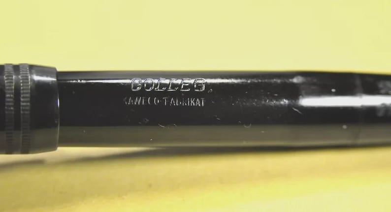 Puede incluir: Una pluma estilogr&aacute;fica negra con acabado brillante que presenta el texto grabado COLLEG y KAWECO-FABRIKAT en el cuerpo. La pluma tiene un cuerpo cil&iacute;ndrico con una secci&oacute;n estriada cerca de la tapa, mostrando letras met&aacute;licas claras.
