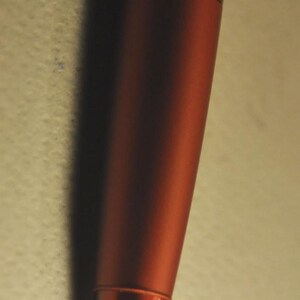 May include: A metallic rust-colored ballpoint pen featuring a smooth, cylindrical barrel and a tapered conical tip. The pen has a consistent matte finish across its body, ending in a pointed metal nib at the very bottom of the writing instrument.