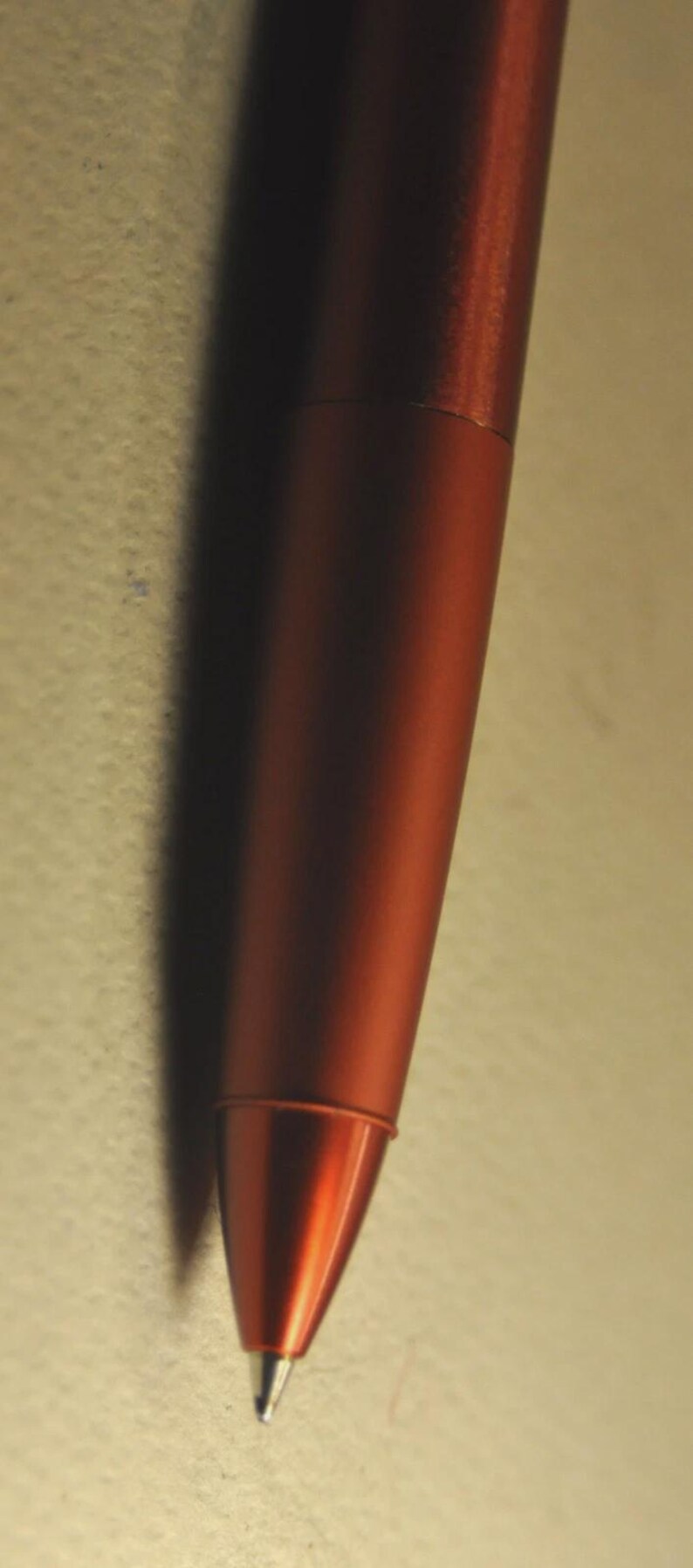 May include: A metallic rust-colored ballpoint pen featuring a smooth, cylindrical barrel and a tapered conical tip. The pen has a consistent matte finish across its body, ending in a pointed metal nib at the very bottom of the writing instrument.