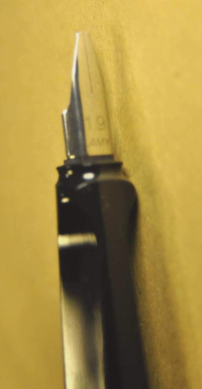 K&ouml;nnte beinhalten: Nahaufnahme eines schwarzen F&uuml;llfederhalters mit silberner Feder. Auf der Feder sind die Zahl "19" und die Buchstaben "AM" eingraviert. Der Stift befindet sich vor einem gelben Hintergrund.