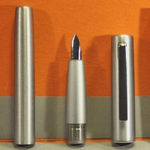 Może przedstawiać: Srebrne pi&oacute;ro wieczne ze stal&oacute;wką ze stali nierdzewnej, rozebrane na części. Widoczne elementy to skuwka, korpus i sekcja z pi&oacute;rem. Pi&oacute;ro jest na pomarańczowym tle.