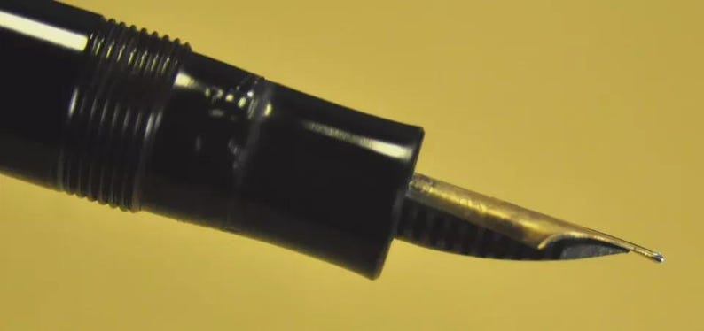 Puede incluir: Una pluma estilogr&aacute;fica negra con un plum&iacute;n de color oro met&aacute;lico. La pluma presenta un cuerpo cil&iacute;ndrico con una secci&oacute;n roscada cerca de la empu&ntilde;adura, que conduce al plum&iacute;n expuesto. El dise&ntilde;o es sencillo y funcional, mostrando la punta.