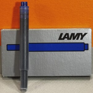 Może przedstawiać: Wkład atramentowy Lamy, wypełniony czarnym tuszem, stoi przed swoim srebrnym pudełkiem. Pudełko ma logo Lamy i niebieski pasek. Wkład ma niebieską nasadkę.