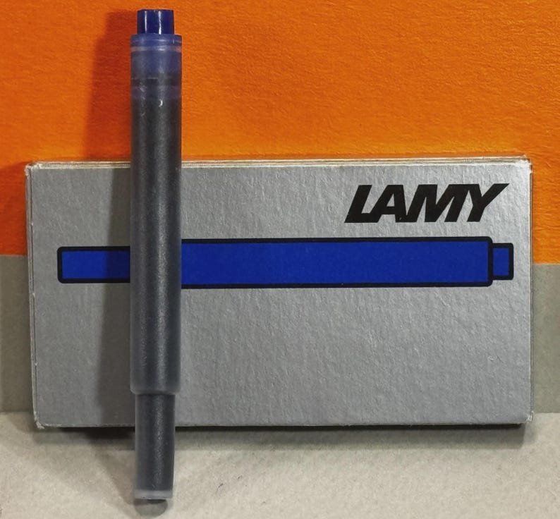 Może przedstawiać: Wkład atramentowy Lamy, wypełniony czarnym tuszem, stoi przed swoim srebrnym pudełkiem. Pudełko ma logo Lamy i niebieski pasek. Wkład ma niebieską nasadkę.