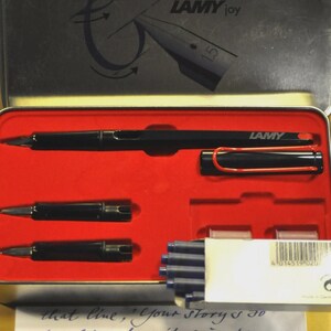 K&ouml;nnte beinhalten: Ein schwarzes Lamy Joy F&uuml;llfederhalter-Set in einem rot ausgekleideten Metallkoffer. Das Set enth&auml;lt den Stift, zwei Federn, Tintenpatronen und einen roten Stiftclip. Der Deckel des Koffers zeigt das Lamy-Logo und den Text "LAMY joy".