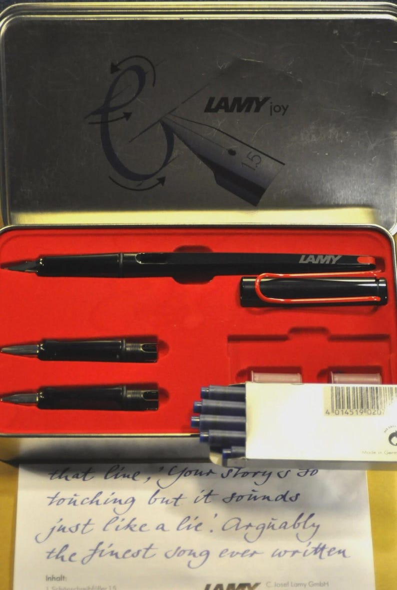 K&ouml;nnte beinhalten: Ein schwarzes Lamy Joy F&uuml;llfederhalter-Set in einem rot ausgekleideten Metallkoffer. Das Set enth&auml;lt den Stift, zwei Federn, Tintenpatronen und einen roten Stiftclip. Der Deckel des Koffers zeigt das Lamy-Logo und den Text "LAMY joy".