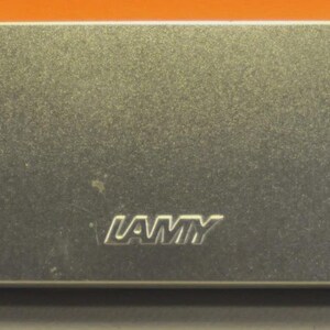 K&ouml;nnte beinhalten: Ein rechteckiges Aufbewahrungsetui aus silberfarbenem Metall mit abgerundeten Ecken. Die Oberfl&auml;che hat ein geb&uuml;rstetes Metall-Finish. Das Wort LAMY ist mittig auf dem Deckel in einer fetten, serifenlosen Schrift eingepr&auml;gt. Das Etui ist geschlossen.