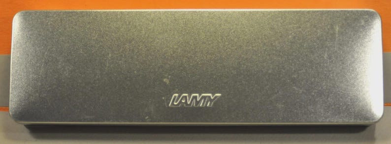 K&ouml;nnte beinhalten: Ein rechteckiges Aufbewahrungsetui aus silberfarbenem Metall mit abgerundeten Ecken. Die Oberfl&auml;che hat ein geb&uuml;rstetes Metall-Finish. Das Wort LAMY ist mittig auf dem Deckel in einer fetten, serifenlosen Schrift eingepr&auml;gt. Das Etui ist geschlossen.