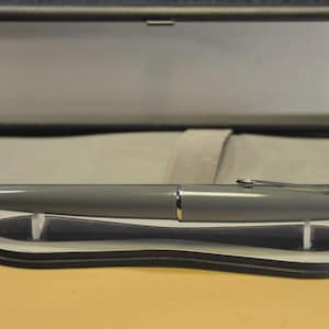 Peut inclure: Un stylo plume gris avec des détails argentés, présenté dans un étui noir ouvert avec un intérieur gris. Le stylo repose sur un présentoir noir et blanc. Le stylo a un design élégant et moderne.