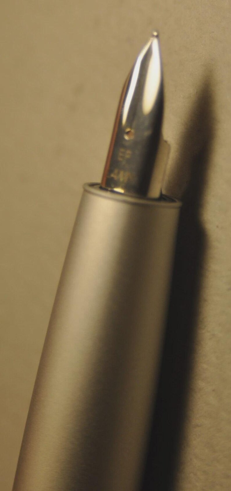 May include: Close-up of a silver fountain pen nib with the text "EF 4M" visible. The pen's body is a matte silver color, and the nib is highly polished, reflecting light. The pen is angled, with a soft background.