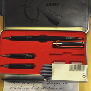 K&ouml;nnte beinhalten: Schwarzes Lamy Joy Stift-Set in einer Metallbox mit rotem Innenraum. Das Set enth&auml;lt einen Stift, zwei Federn und blaue Tintenpatronen. Der Deckel zeigt das Lamy Joy Logo und eine Stiftillustration.