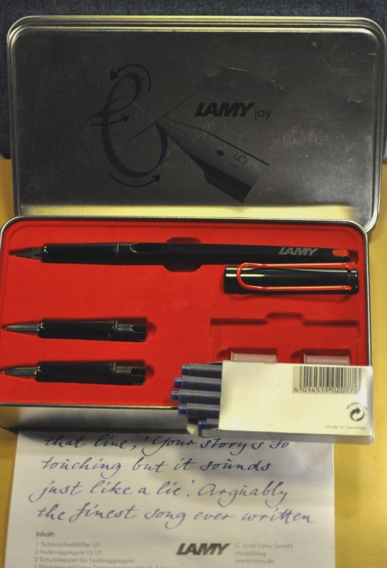 K&ouml;nnte beinhalten: Schwarzes Lamy Joy Stift-Set in einer Metallbox mit rotem Innenraum. Das Set enth&auml;lt einen Stift, zwei Federn und blaue Tintenpatronen. Der Deckel zeigt das Lamy Joy Logo und eine Stiftillustration.