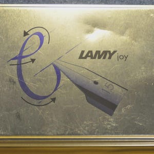 K&ouml;nnte beinhalten: Rechteckige, silberfarbene Metallbox mit den Worten "LAMY joy" und einem stilisierten Federdesign. Das Design beinhaltet eine lila Wirbel mit Pfeilen. Die Box hat eine geb&uuml;rstete Metallstruktur.