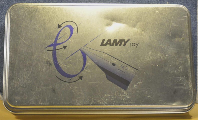 K&ouml;nnte beinhalten: Rechteckige, silberfarbene Metallbox mit den Worten "LAMY joy" und einem stilisierten Federdesign. Das Design beinhaltet eine lila Wirbel mit Pfeilen. Die Box hat eine geb&uuml;rstete Metallstruktur.