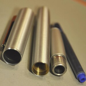 Może przedstawiać: Rozłożony srebrny długopis z niebieskim wkładem. Elementy długopisu obejmują nasadkę z klipsem, korpus i gwintowaną końc&oacute;wkę. Wnętrze korpusu długopisu ma złoty kolor.