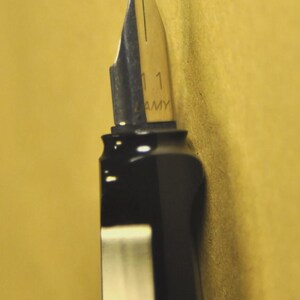 K&ouml;nnte beinhalten: Nahaufnahme eines schwarzen F&uuml;llfederhalters mit silberner Feder. Auf der Feder steht "1.1 AMY". Der Stift befindet sich vor einem gelben Hintergrund.