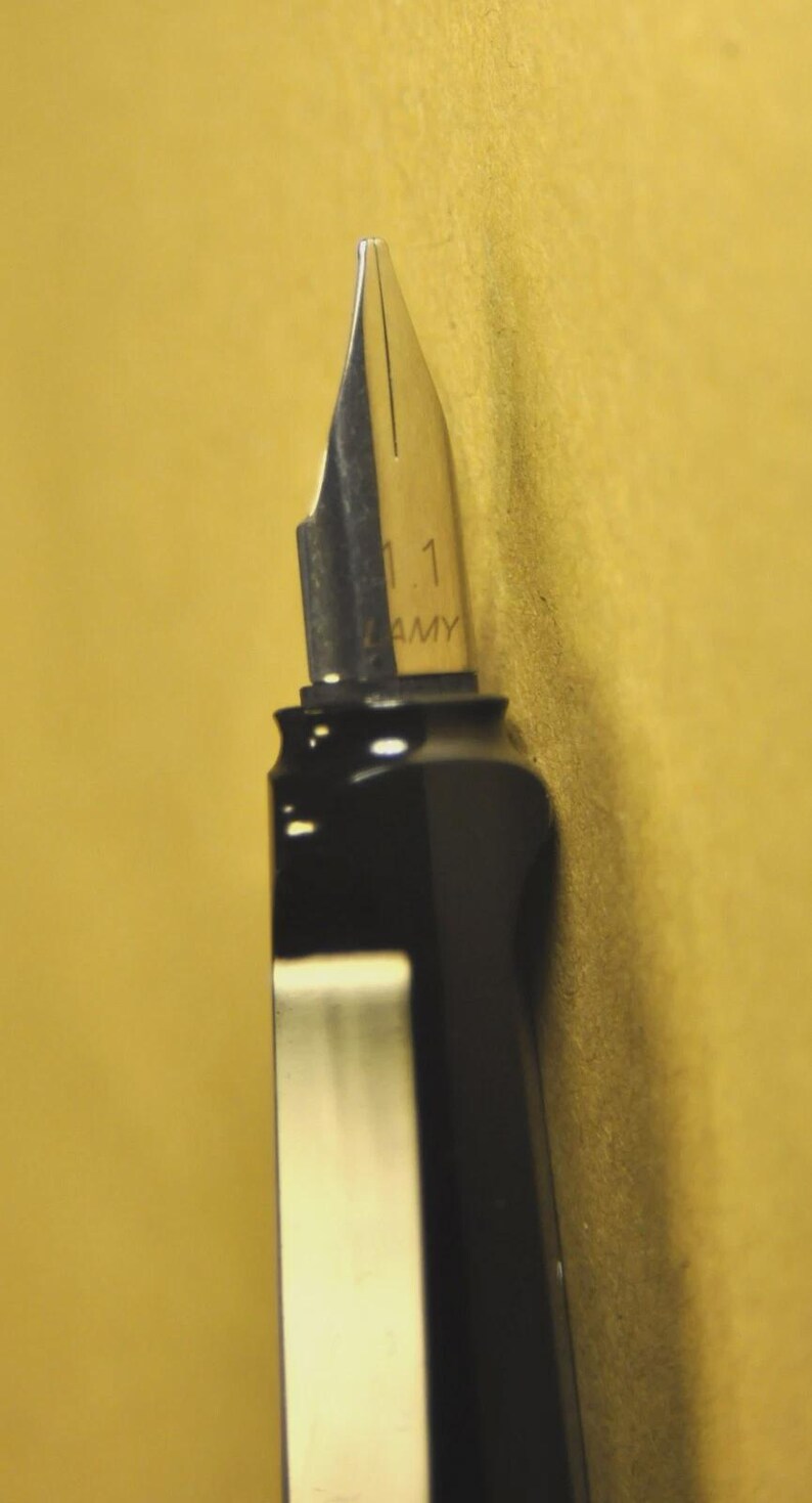 K&ouml;nnte beinhalten: Nahaufnahme eines schwarzen F&uuml;llfederhalters mit silberner Feder. Auf der Feder steht "1.1 AMY". Der Stift befindet sich vor einem gelben Hintergrund.